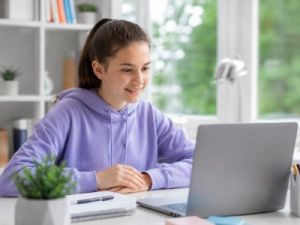 Middle school girl participating in an online tutoring session, smiling optimistically while looking at her laptop at a bright study desk with notebooks and school supplies in a modern home learning environment.